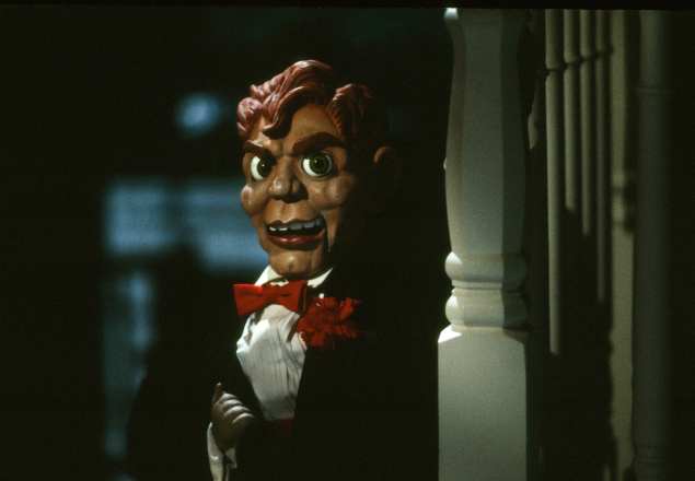 A plastic ventriloquist dummy peeks out from behind a staircase. He is wearing a tuxedo and has brown colored hair.